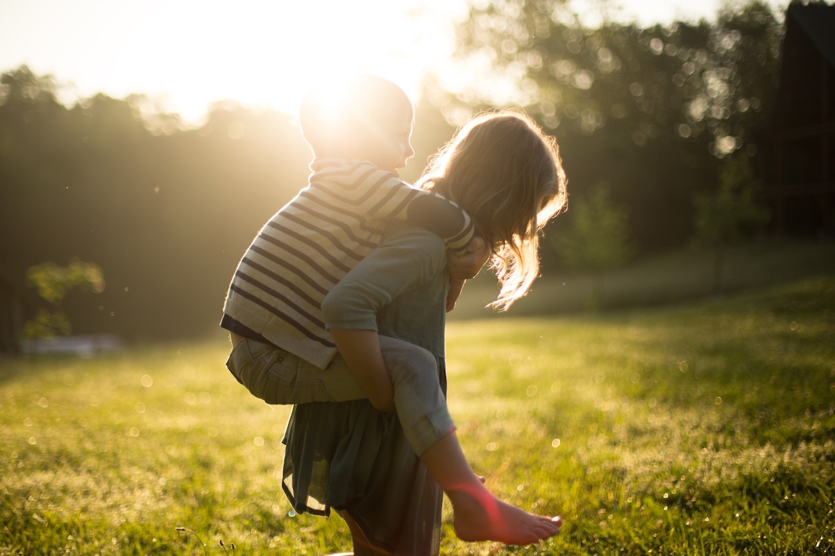 Older child giving piggy back ride to younger child in park at sunset