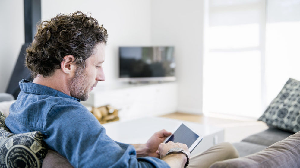 Middle aged man sitting on couch in work clothes, looking at a tablet device