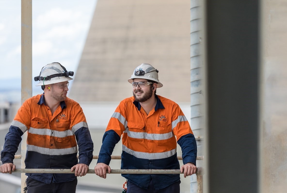 Two men in hi-vis hardhats talking with the cooling towers in the background.